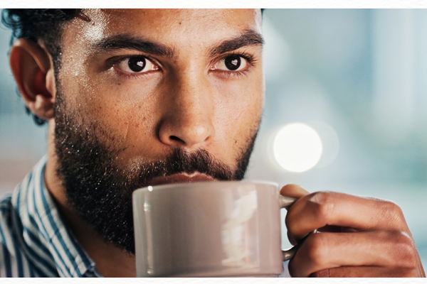 A close-up of a bearded man wearing a striped shirt, holding a white mug with both hands and sipping from it. His expression is thoughtful, and bright natural light softly blurs the background.