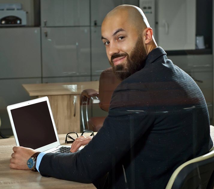 A bearded man with a shaved head, wearing a dark jacket and blue watch, sits at a desk with an open laptop. He looks over his shoulder and smiles at the camera. The background shows office furniture and a glass cabinet.