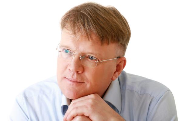 A middle-aged man with light skin, short blonde hair, and glasses looks thoughtfully to the side. He rests his chin on his hands and wears a light blue collared shirt. The background is plain white.